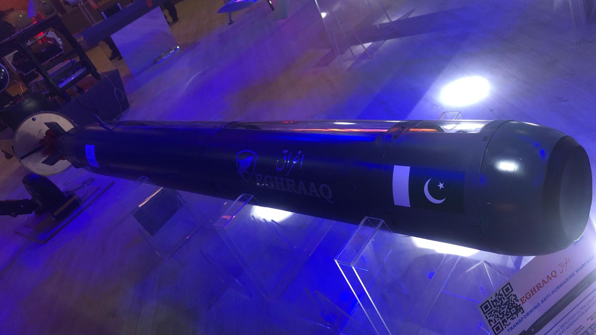 The GIDS Eghraaq, an ultra-lightweight anti-submarine warfare torpedo, showcased on a transparent stand under blue lighting. The torpedo features sleek black casing with the Pakistan flag and the Eghraaq logo prominently displayed, symbolizing advanced indigenous technology. The setting highlights its modern design and engineering, with informational signage visible in the background.