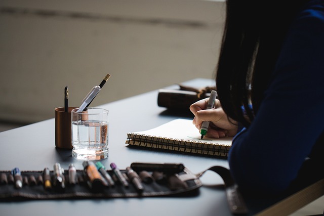 Person writing in a notebook on a desk with art supplies, pens, and a glass of water, creating a calm and focused study environment.