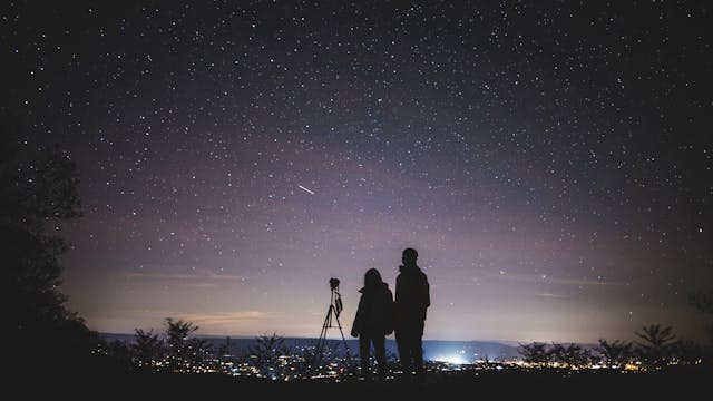 Two people silhouetted against a brilliant starry night sky, standing next to a telescope on a hill overlooking a glowing cityscape in the distance.