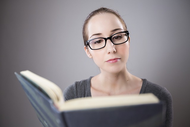 A woman wearing black-framed glasses is reading a book attentively. She has a focused expression, and the background is softly blurred, emphasizing her engagement with the book. She is wearing a gray top, creating a neutral and calm setting.