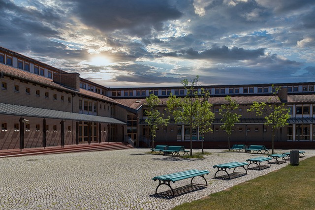 Courtyard of a modern school building with benches, trees, and a cloudy sky at sunset.