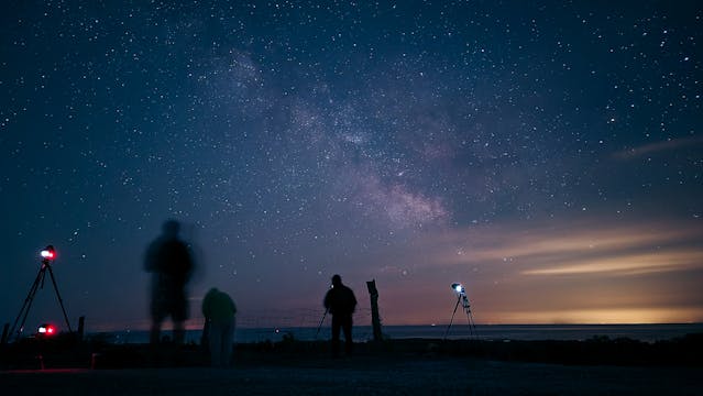 Silhouettes of people stargazing under a vibrant night sky filled with stars and the Milky Way, accompanied by telescopes on a quiet observation site near the horizon.