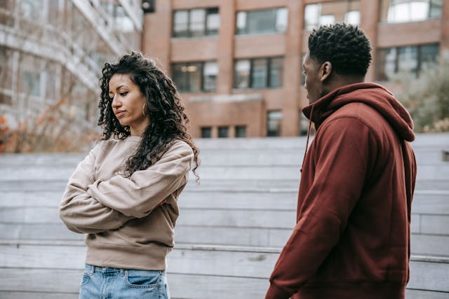 A woman with her arms crossed looking away, appearing upset, while a man in a hoodie stands nearby trying to engage her in conversation. The image reflects the dynamics of the silent treatment in a relationship.