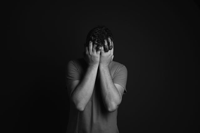 A black-and-white photograph of a person standing with their face buried in their hands, expressing emotions such as distress, sadness, or frustration, set against a dark background.